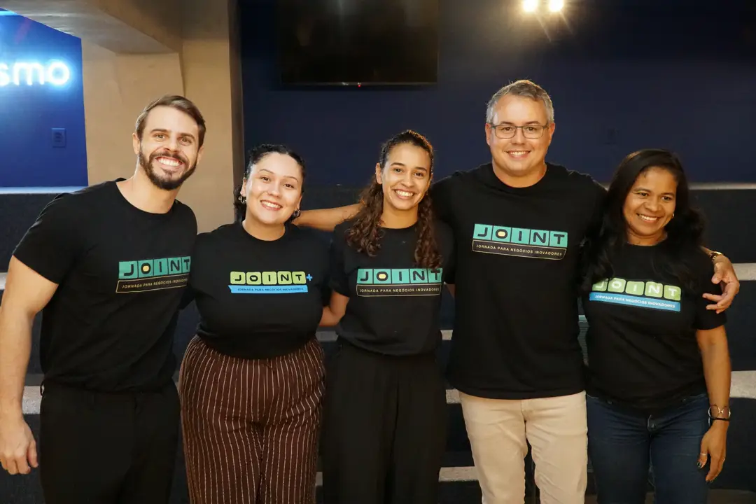 Cinco pessoas da equipe Joint posam sorrindo lado a lado, com camisetas do programa.
