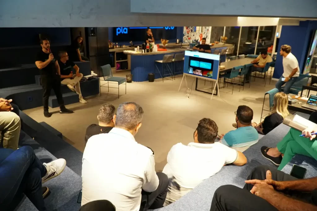 Apresentação em tela para participantes na área em estilo lounge da Ampla Comunicação, com arquibancadas azuis e mesas ao fundo.