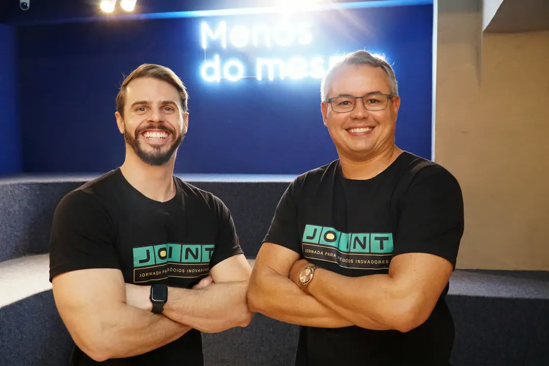 Gabriel Melo e Flammarion Cysneiros sorrindo, usando camisetas da Joint, em ambiente corporativo.