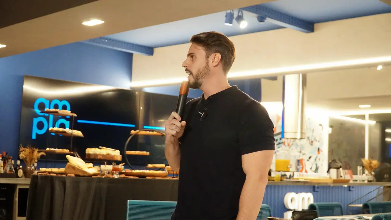 Gabriel Melo com microfone fala em pé durante evento em espaço corporativo.