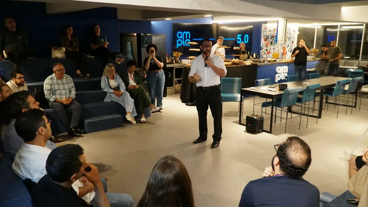 Leonardo Estevam fala ao microfone diante de plateia sentada em evento, com área de buffet e participantes ao fundo.